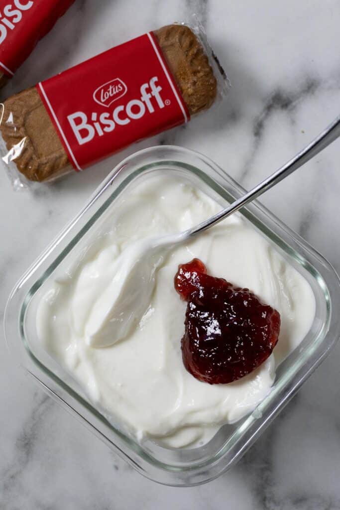 A glass dish of creamy yogurt topped with a spoonful of red jam, with a spoon resting inside. Two packaged Lotus Biscoff biscuits are placed nearby on a marble surface.