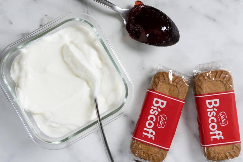 A glass container of plain yogurt with a spoon, a spoonful of dark jam, and two wrapped Lotus Biscoff cookies on a white marble surface.