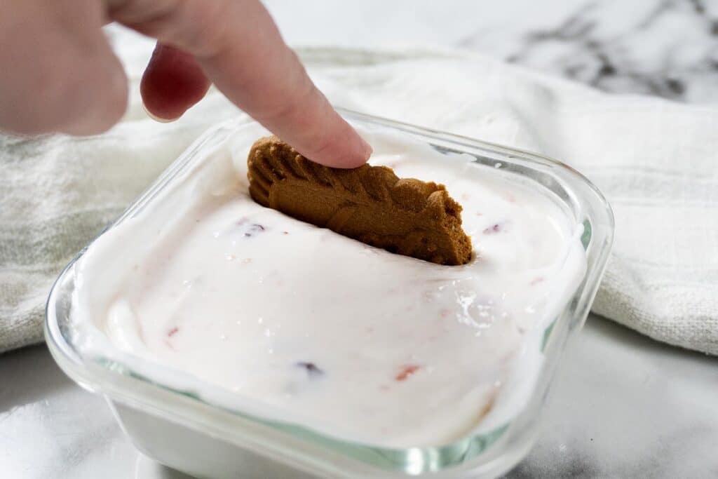 A hand dips a brown cookie into a square glass dish filled with creamy yogurt that contains small pieces of fruit, on a marble surface with a white cloth in the background.