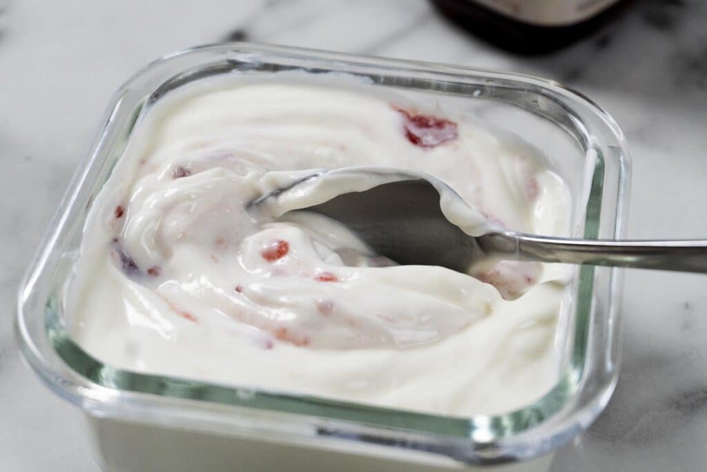 A close-up of a spoon stirring creamy yogurt with visible swirls of strawberry preserves in a glass container on a white surface.