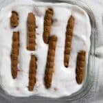 A glass dish filled with a creamy white dessert topped with rows of rectangular brown biscuits, set on a white marble surface with a light textured cloth nearby.