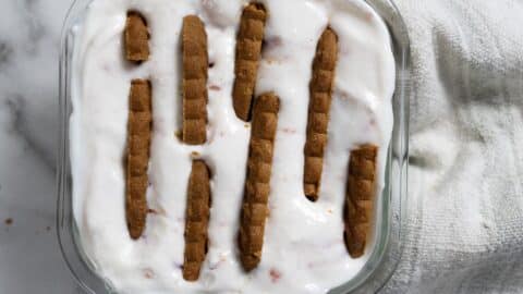 A glass dish filled with a creamy white dessert topped with rows of rectangular brown biscuits, set on a white marble surface with a light textured cloth nearby.