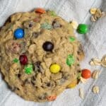 A large oatmeal cookie with colorful chocolate candies and chocolate chips rests on a light-colored cloth, with a few oats and candies scattered nearby.
