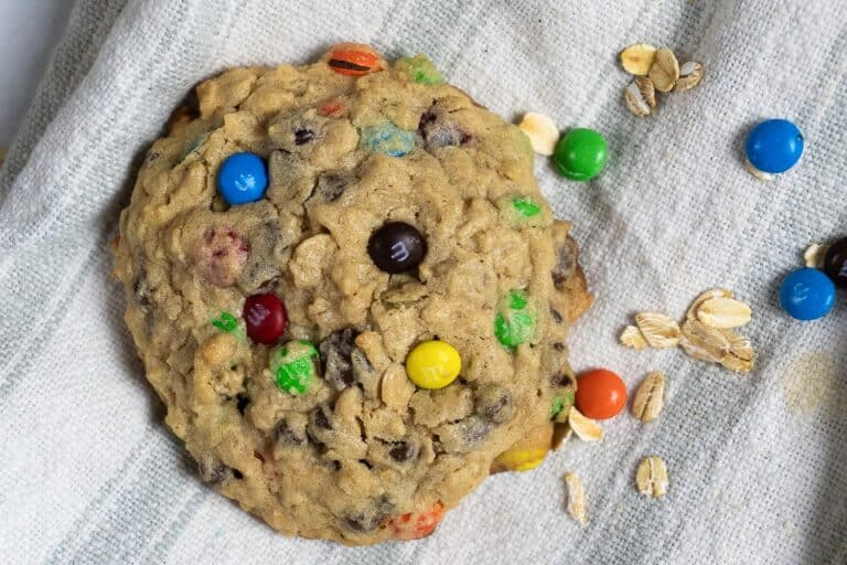 A large oatmeal cookie with colorful chocolate candies and chocolate chips rests on a light-colored cloth, with a few oats and candies scattered nearby.