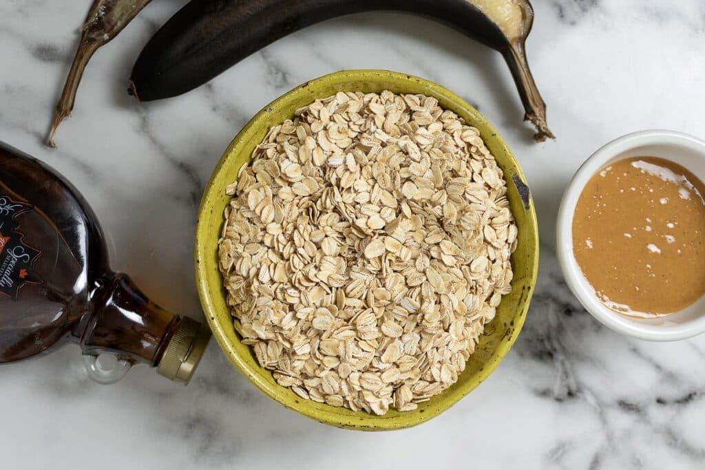 A yellow bowl filled with rolled oats sits on a marble surface, surrounded by a dark overripe banana, a bottle of maple syrup, and a small white bowl of peanut butter.
