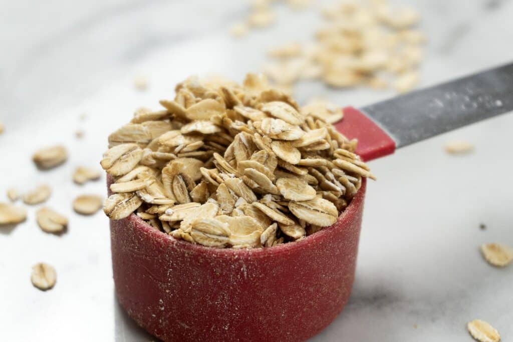 A red measuring cup filled with rolled oats, with additional oats scattered on a white marble surface around it.