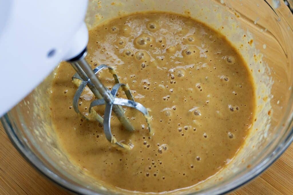 A close-up of a glass bowl filled with light brown batter being mixed with a hand mixer on a wooden surface. Bubbles are visible on the surface of the batter.