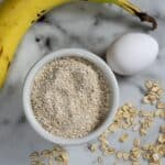 A ripe banana, a white egg, a bowl filled with oat flour, and scattered oats are arranged on a white marble surface.