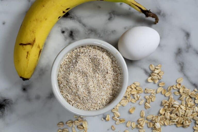 A ripe banana, a white egg, a bowl filled with oat flour, and scattered oats are arranged on a white marble surface.