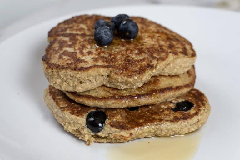 Three golden-brown pancakes stacked on a white plate, topped with a few fresh blueberries and drizzled with syrup. More blueberries are visible within the pancakes.