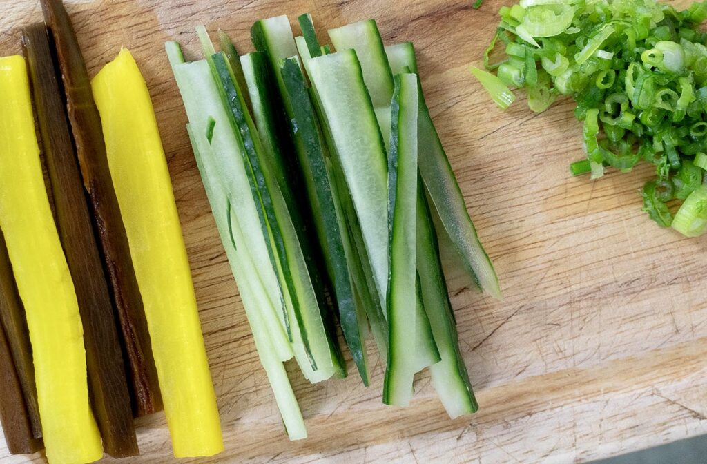 Sliced yellow and brown pickled radish, cucumber sticks, and chopped green onions are arranged on a wooden cutting board.