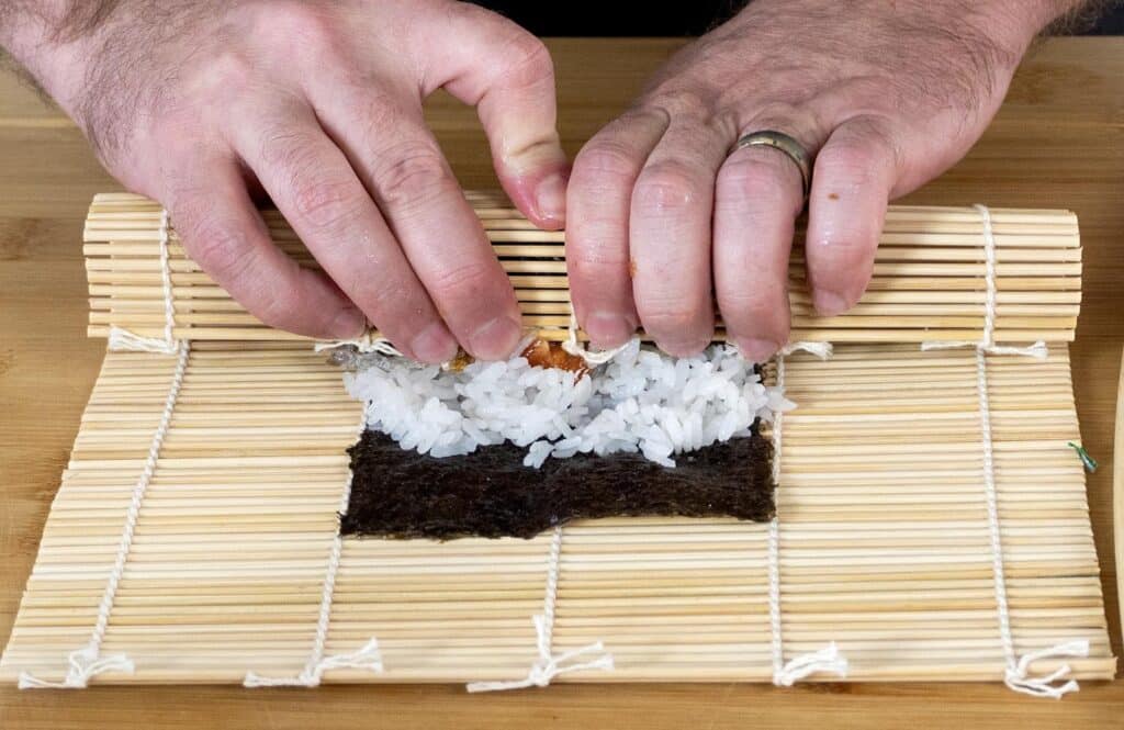Close-up of hands rolling sushi with a bamboo mat, spreading rice and filling on a sheet of nori seaweed, on a wooden surface.