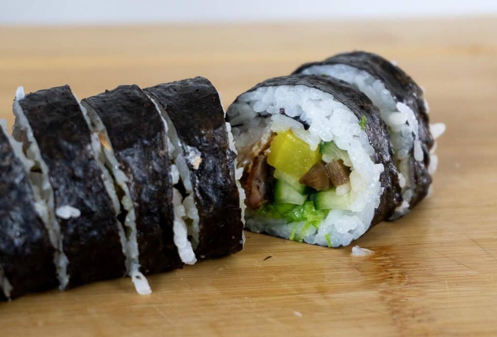 Close-up of sliced sushi rolls on a wooden surface, showing white rice, cucumber, yellow pickled radish, and other vegetables wrapped in seaweed.