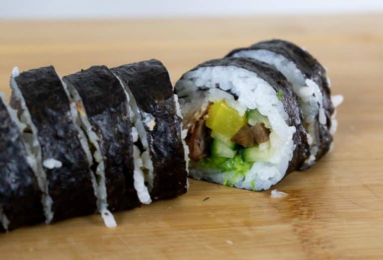 Close-up of sliced sushi rolls on a wooden surface, showing white rice, cucumber, yellow pickled radish, and other vegetables wrapped in seaweed.