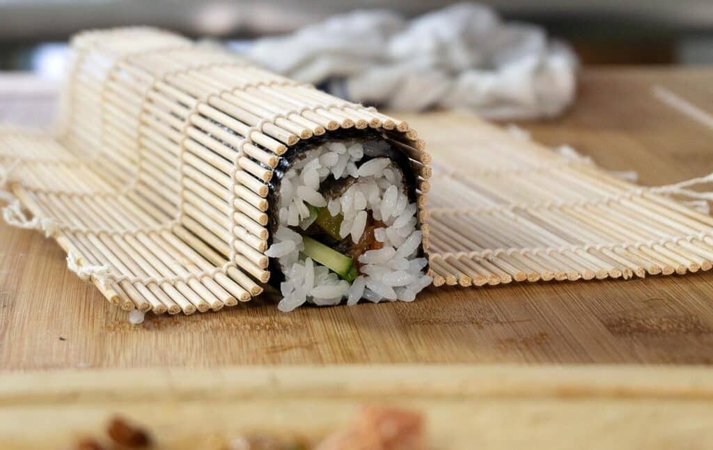 A sushi roll filled with rice and vegetables is partially wrapped in a bamboo sushi mat on a wooden surface, ready to be sliced.