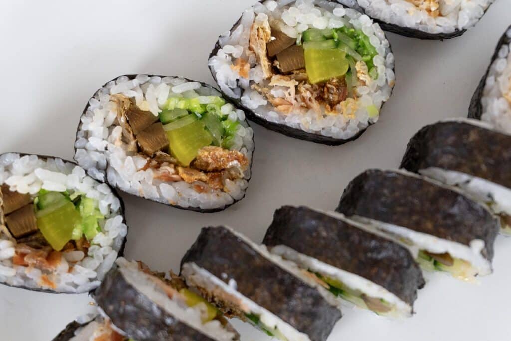 Close-up of sliced sushi rolls filled with rice, yellow pickled radish, green vegetables, and other ingredients, arranged in a diagonal line on a white surface.
