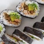 Close-up of sliced sushi rolls filled with rice, yellow pickled radish, green vegetables, and other ingredients, arranged in a diagonal line on a white surface.