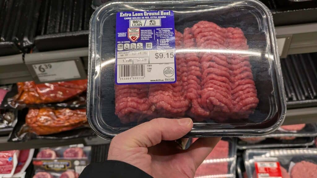 A hand holds a package of extra lean ground beef priced at $9.15 in a grocery store meat section, highlighting current ground beef prices. The beef is pink and neatly packed in rows inside a clear plastic container with a black tray.
