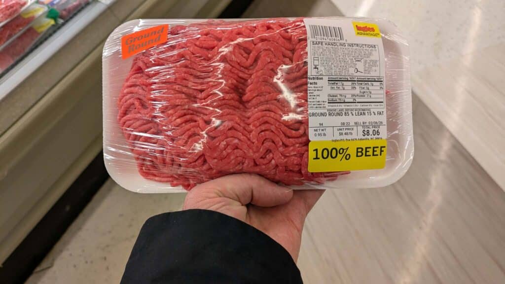 A hand holds a package of ground beef labeled 100% BEEF, priced at $8.06. Wrapped in clear plastic, the label details weight, handling instructions, and highlights current ground beef prices.