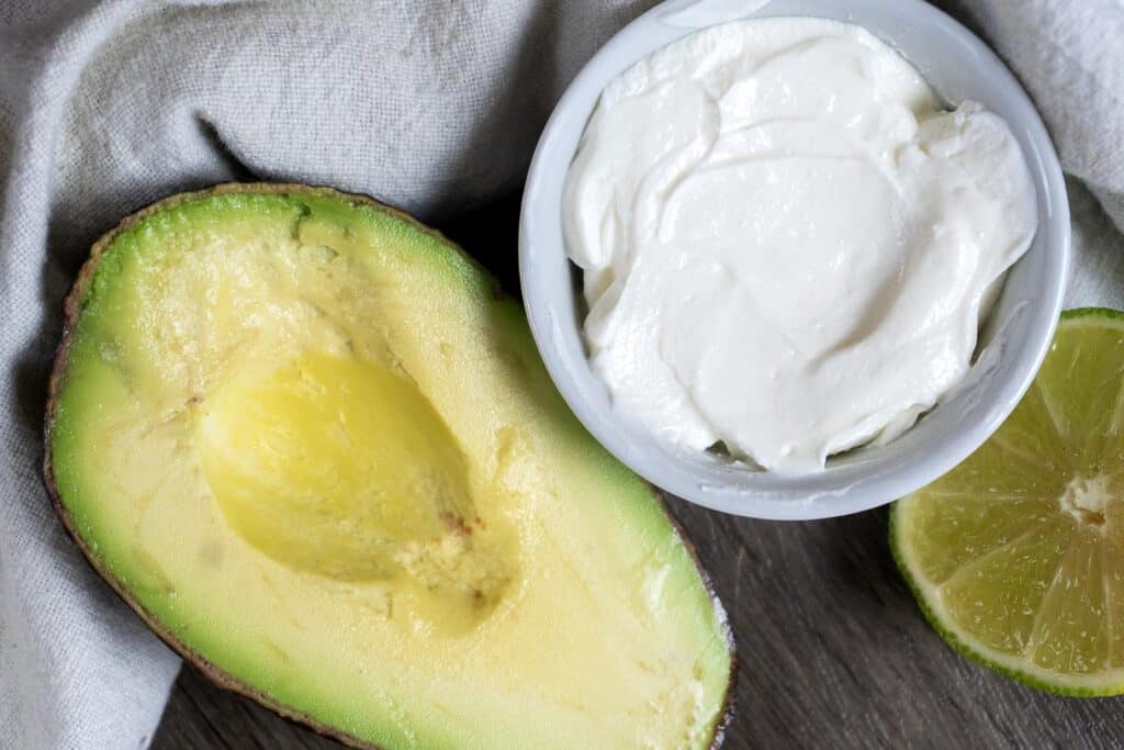 A halved avocado, a bowl of creamy white sauce, and a halved lime are arranged on a wooden surface with a light gray cloth beside them.