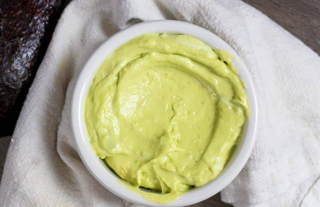 A white bowl filled with smooth, creamy avocado sauce sits on a white cloth napkin, with part of a dark avocado skin visible to the side.