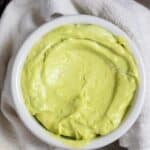 A white bowl filled with smooth, creamy avocado sauce sits on a white cloth napkin, with part of a dark avocado skin visible to the side.
