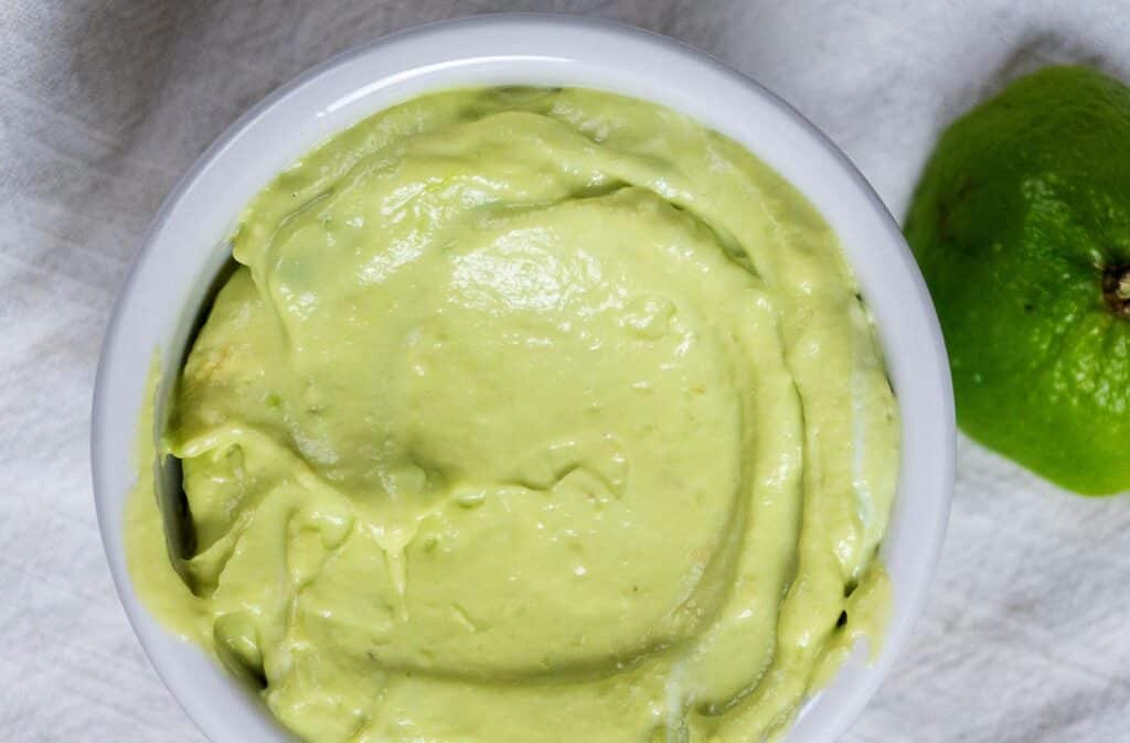 A close-up of a white bowl filled with creamy, smooth avocado dip on a light-colored cloth, with a whole lime next to it.