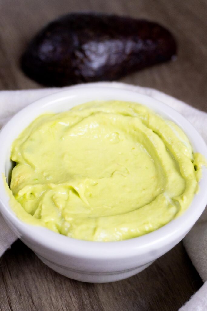 A small white bowl filled with creamy, light green avocado spread sits on a wooden surface, with a dark avocado skin visible in the background.