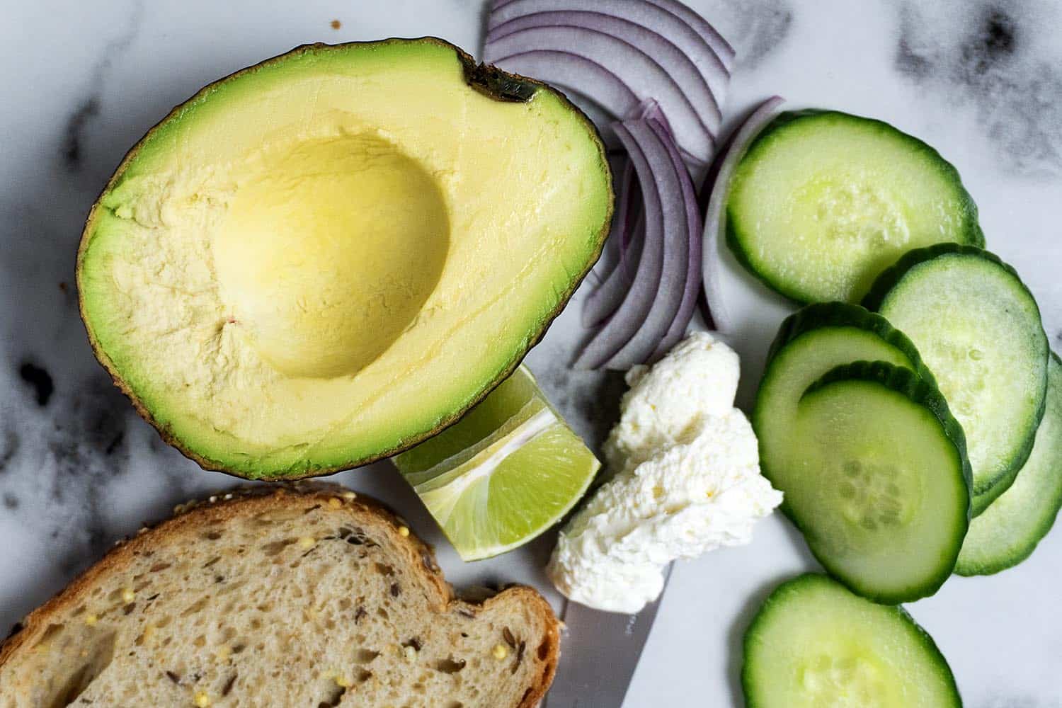 A halved avocado, sliced cucumber, red onion, a wedge of lime, ricotta cheese, and a slice of whole grain bread are arranged on a marble surface.