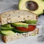 A sandwich with whole grain bread, cucumber slices, tomato, red onion, avocado, and a spread, sits on a marble surface. A halved avocado is in the background.