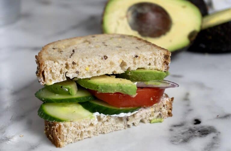 A sandwich with whole grain bread, cucumber slices, tomato, red onion, avocado, and a spread, sits on a marble surface. A halved avocado is in the background.