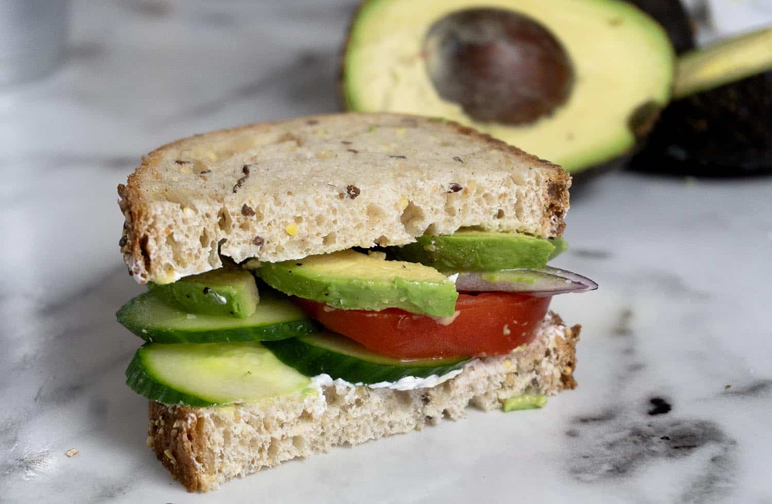 A sandwich with whole grain bread, cucumber slices, tomato, red onion, avocado, and a spread, sits on a marble surface. A halved avocado is in the background.