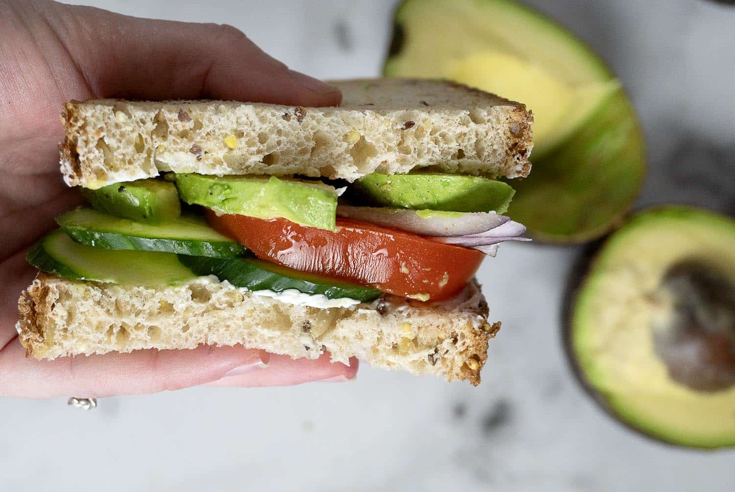 A hand holding a sandwich with visible layers of sliced avocado, tomato, cucumber, and red onion between two pieces of multigrain bread. In the background, there are halved avocados on a light surface.