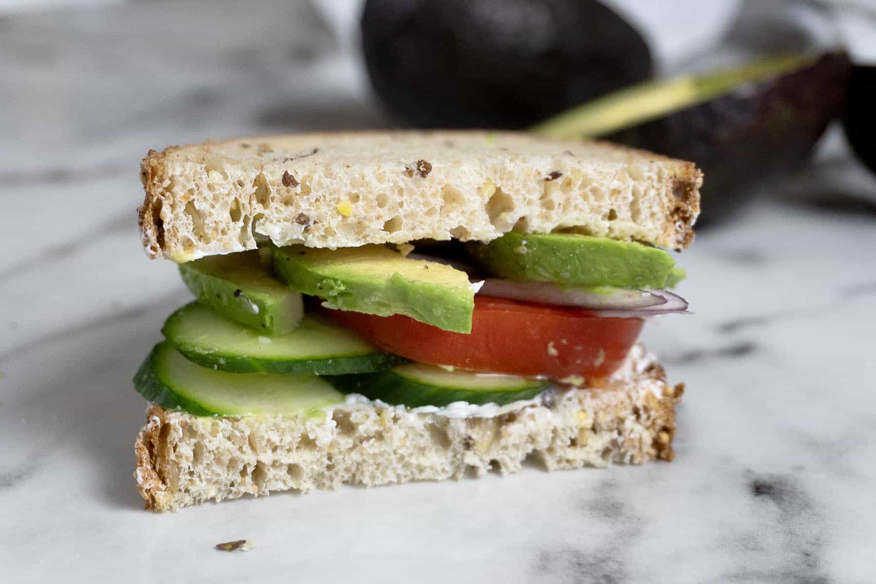 A close-up of a sandwich on whole grain bread, filled with slices of avocado, tomato, cucumber, and red onion, with a light spread, on a marble surface. An avocado is blurred in the background.