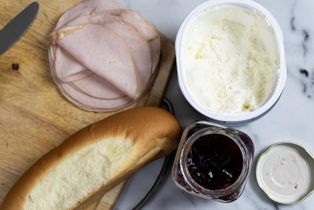 Sliced deli meat, a hot dog bun, a tub of cream cheese, and an open jar of grape jelly are arranged on a wooden cutting board and marble surface.