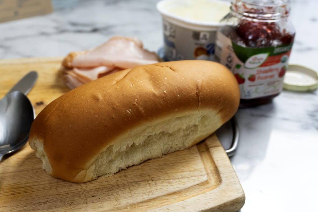 A plain sandwich roll sits on a wooden cutting board next to a spoon, deli turkey slices, a container of spread, and an open jar of strawberry preserves on a marble countertop.
