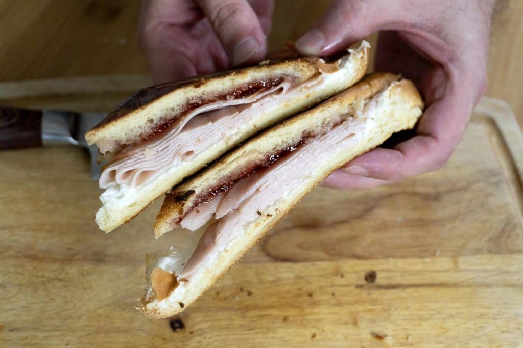 A person holds two halves of a sandwich filled with sliced turkey and red jam, showing the layers inside. The sandwich is on a wooden cutting board with a knife nearby.