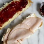 An open sandwich on a marble surface: one slice of bread with cream cheese and turkey slices, the other with strawberry jam. A spoon with jam rests nearby.