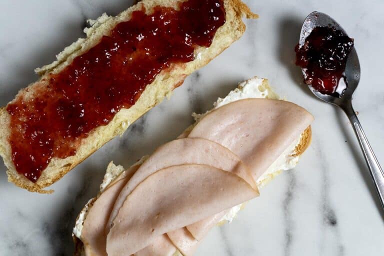 An open sandwich on a marble surface: one slice of bread with cream cheese and turkey slices, the other with strawberry jam. A spoon with jam rests nearby.