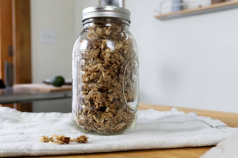 A glass mason jar filled with granola sits on a wooden table covered with a white cloth. A few pieces of granola are scattered near the jar. A blurred kitchen background is visible.