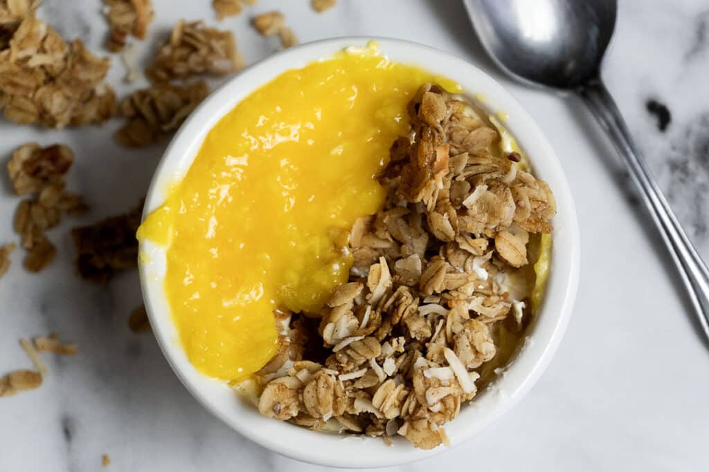 A white bowl filled with granola and bright yellow yogurt or fruit puree, sitting on a marble surface with a spoon nearby and some granola pieces scattered around.