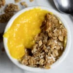 A white bowl filled with granola and bright yellow yogurt or fruit puree, sitting on a marble surface with a spoon nearby and some granola pieces scattered around.