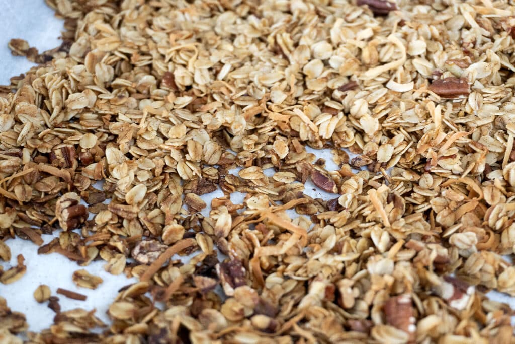 A close-up of homemade granola with toasted oats, coconut flakes, and chopped nuts spread on parchment paper.