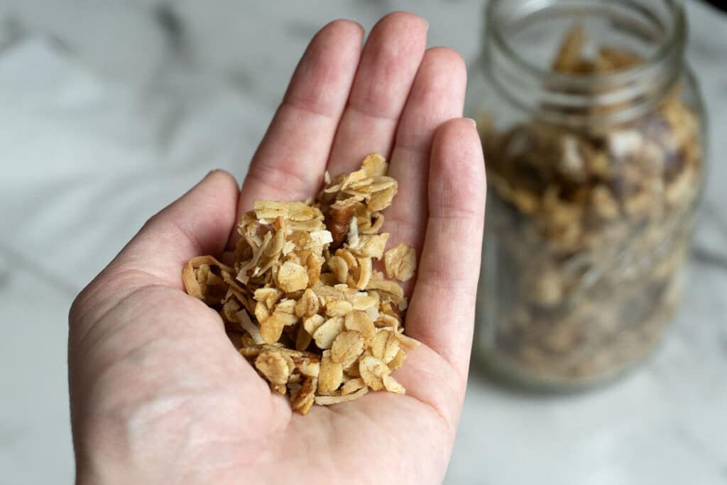 A hand holding a small handful of granola, with a glass jar of granola in the blurred background on a marble surface.