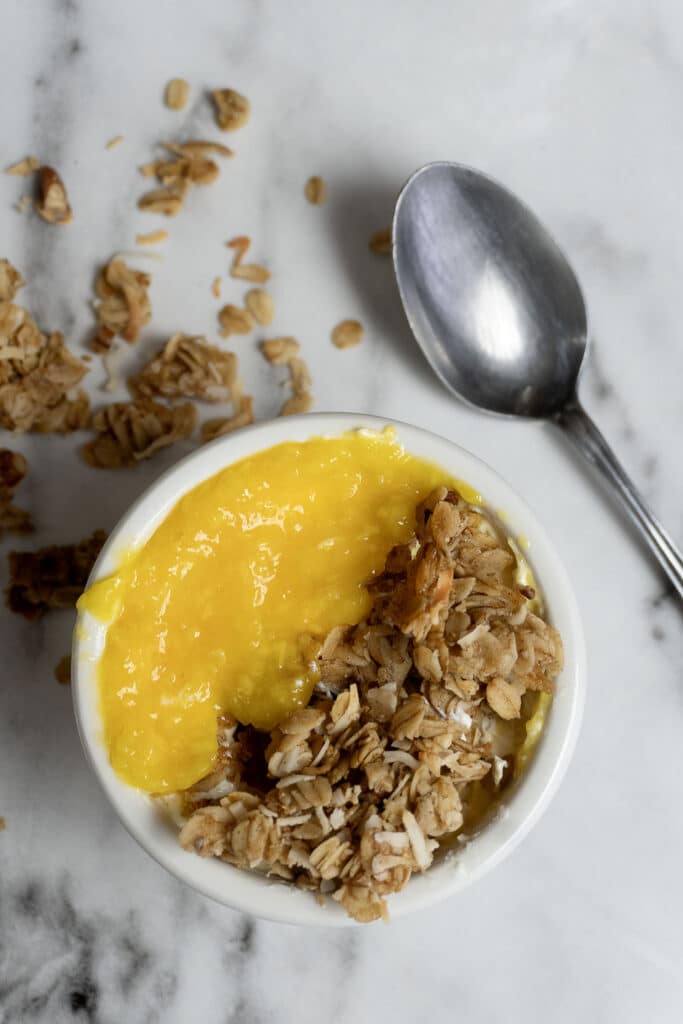 A small white bowl filled with yellow mango puree and granola sits on a marble surface, with a spoon and some scattered granola pieces nearby.