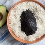 A whole avocado with dark skin sits in a bowl of uncooked white rice, next to a halved avocado on a white towel with blue stripes.