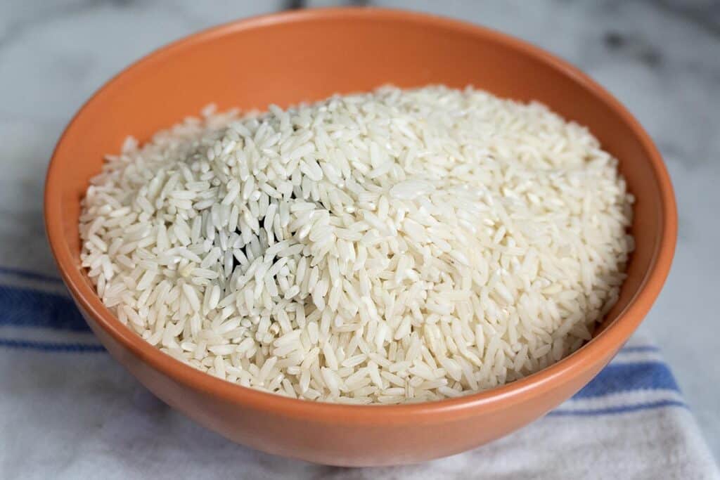 A full orange bowl filled with uncooked white rice sits on a blue and white striped cloth on a marble surface.