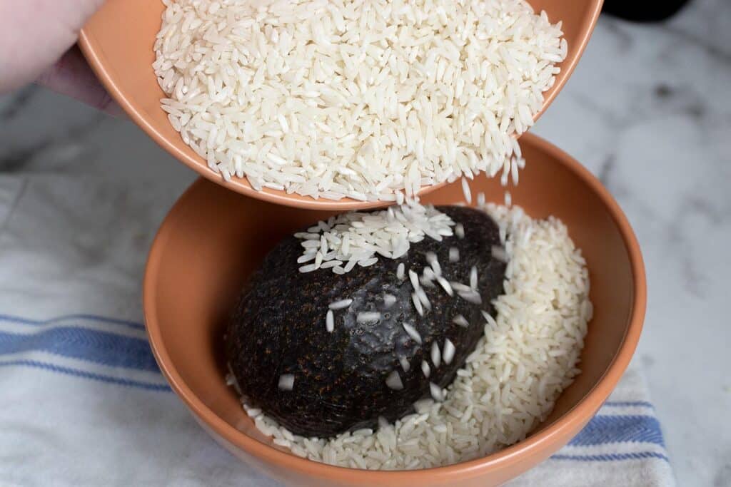 A hand pours uncooked white rice from a bowl over a dark avocado placed in another bowl, with a blue-striped cloth underneath on a marble surface.