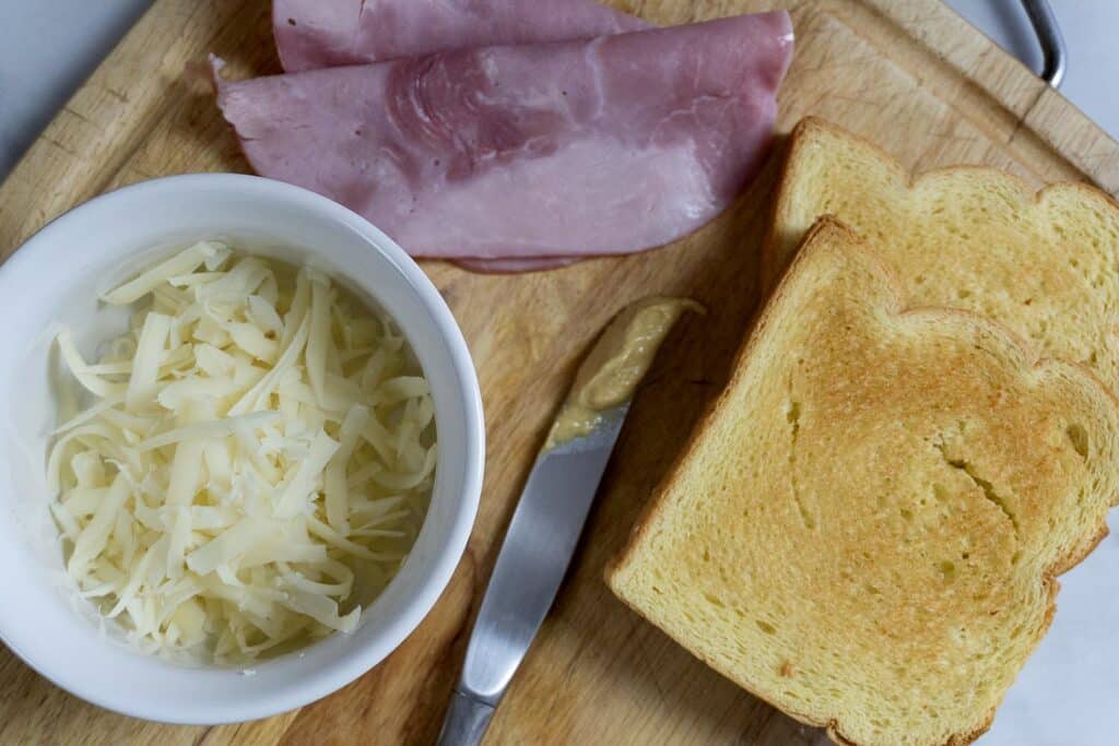 A cutting board with two slices of toasted bread, two slices of ham, a bowl of shredded cheese, a knife, and a small amount of mustard.