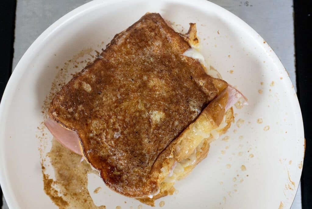 A cooked ham and cheese sandwich with golden-brown, crispy bread sits on a white plate, showing some melted cheese and ham at the edges.